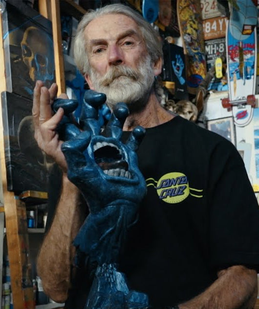 Jim Philips holding a sculpture of the Santa Cruz screaming hand icon, in an artists studio with shelving lined with paint.