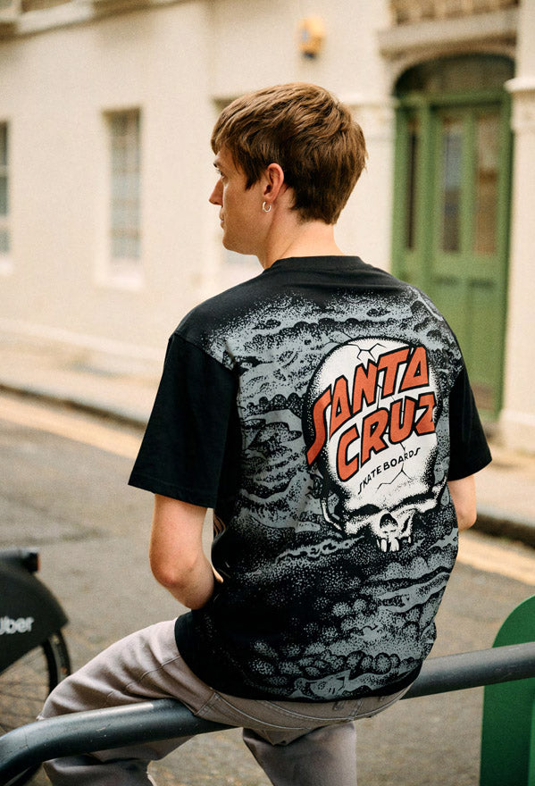 Man wearing a black Santa Cruz t-shirt with a 'Santa Cruz' logo and reaper print sitting on a railing outdoors.