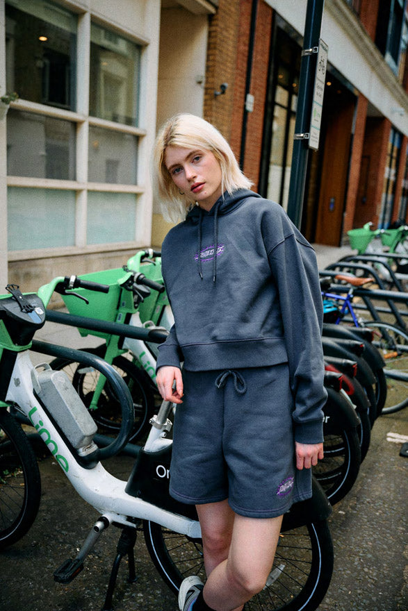 Woman wearing a slate Santa Cruz Acid Oval hoodie and shorts with a logo in an urban setting with bicycles.