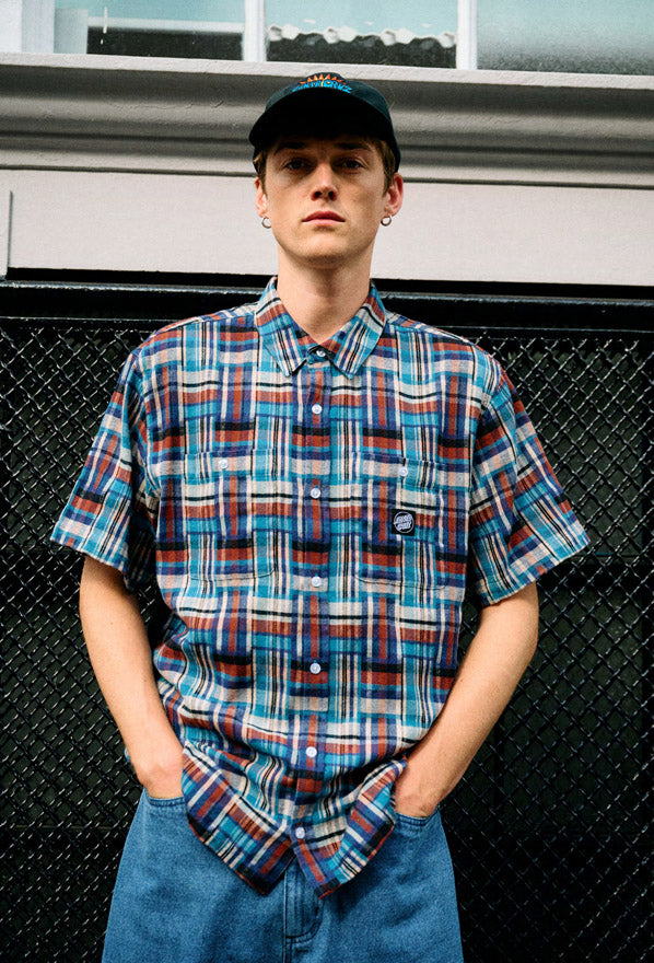 Person wearing a Santa Cruz Milo check shirt and cap standing against a black metal fence.