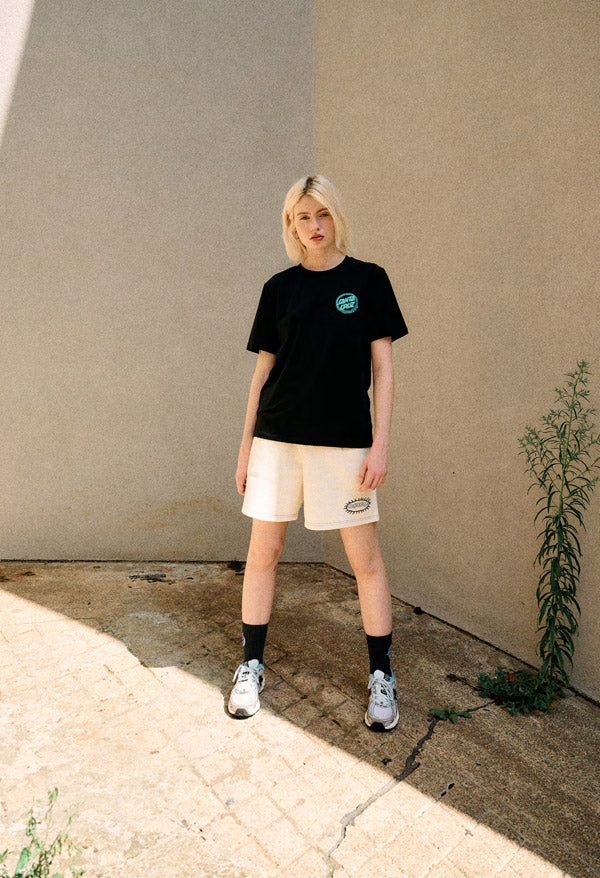 Woman wearing a black Santa Cruz Dizzy Dot t-shirt and beige shorts standing against a plain wall.