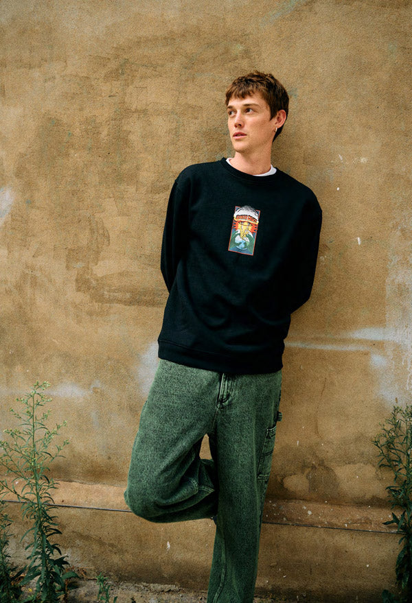 Person wearing a black Santa Cruz EOTW sweatshirt with a graphic design and green jeans against a textured beige wall.