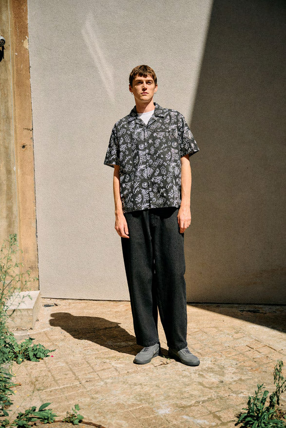 Man wearing a Santa Cruz Finn scribble pattern shirt and black Kayce pants standing against a plain wall.