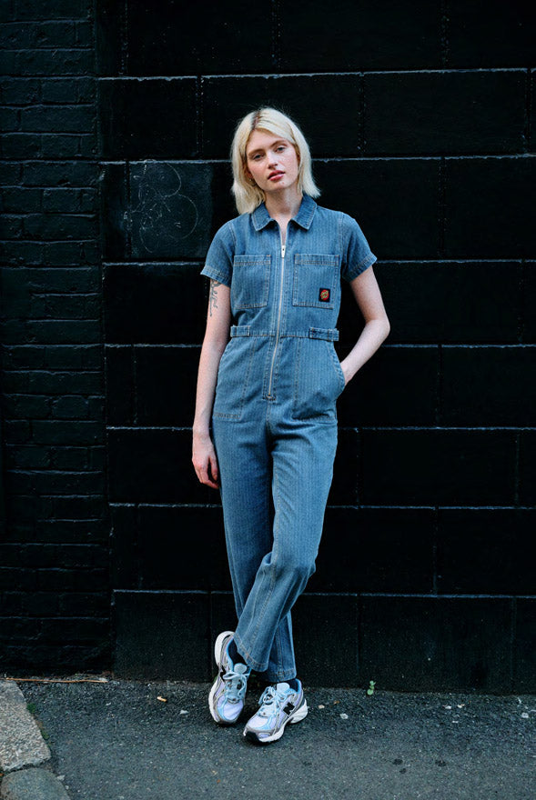 Woman wearing a light blue stripe Santa Cruz Lune Up Utility jumpsuit standing against a black brick wall.