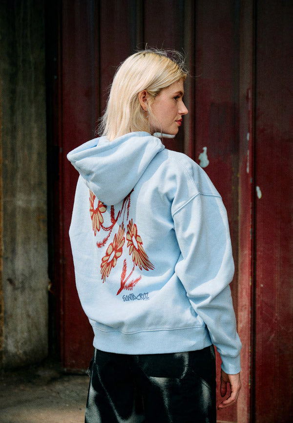 Woman wearing a light blue Santa Cruz zip hoodie with a red floral design on the back, standing in front of a red door