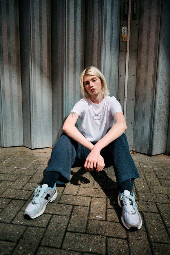 Woman sitting on a brick pavement wearing a white Santa Cruz Acid Oval t-shirt against a metallic wall