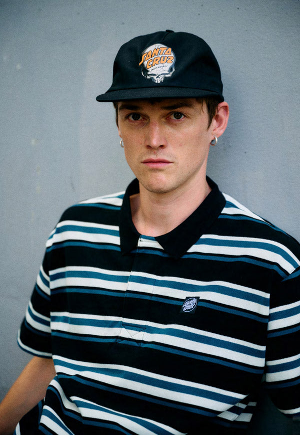 Person wearing a black and teal striped Santa Cruz Cera polo shirt with a cap against a gray background