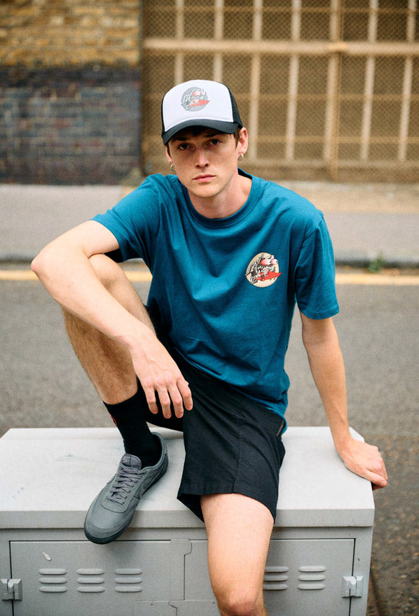 Man wearing a dark teal Santa Cruz Meek Slasher Box t-shirt and black shorts sitting on a locker with a cap.
