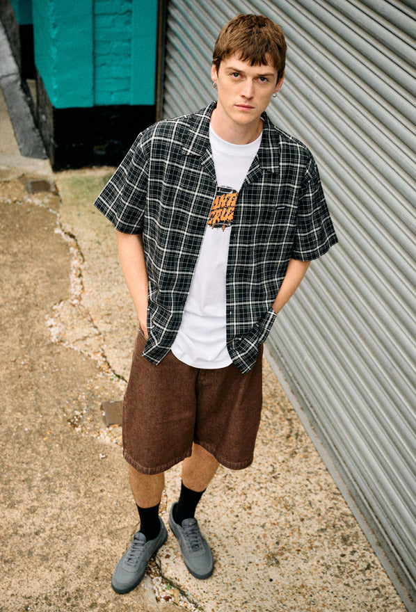 Man wearing a Santa Cruz Flint check shirt over a white graphic t-shirt with brown shorts and gray shoes, standing against a corrugated metal wall.