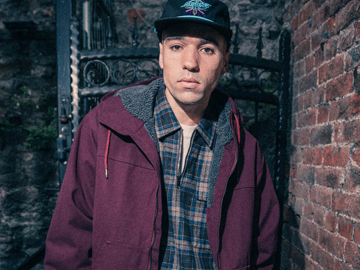 Santa Cruz Men's Clothing. A man wearing a burgundy hooded jacket with a gray fleece lining, a blue and beige plaid shirt, and a black cap with a green graphic, standing in front of a brick wall and black metal gate.