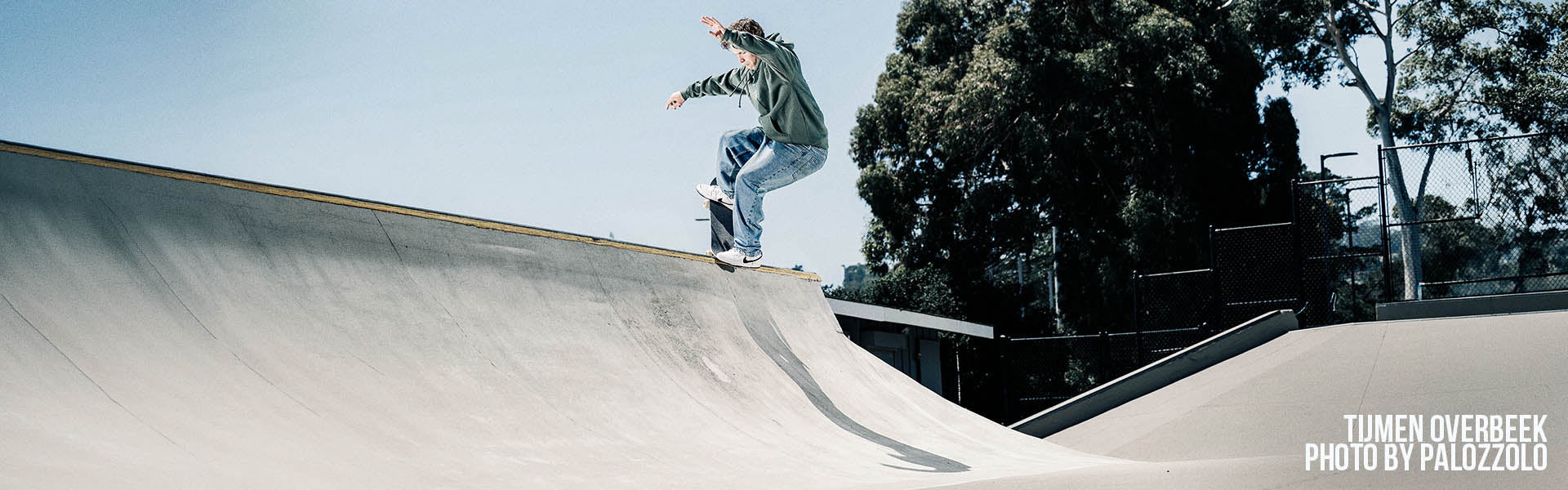 Santa Cruz Skateboards. A skateboarder in a green jacket and light blue jeans rides along the curved edge of a concrete skatepark ramp under a clear blue sky, with trees in the background. 