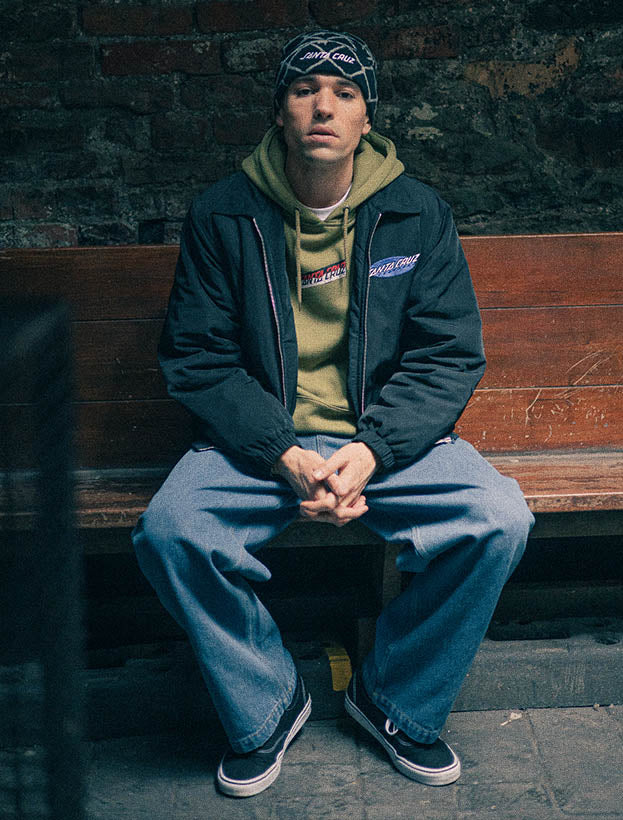 A man sits on a wooden bench against a dark brick wall, wearing a black Santa Cruz jacket over a green hoodie, with blue denim jeans.