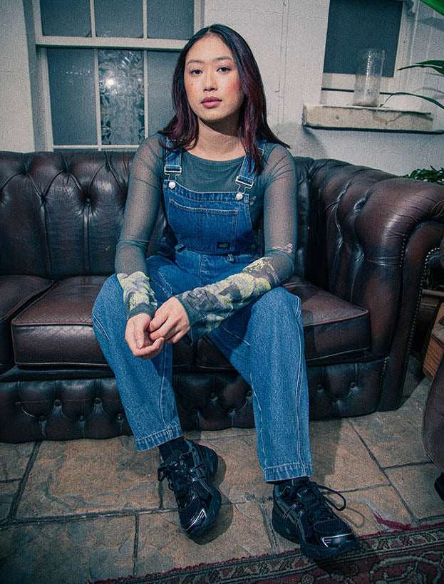 A women sits on a brown sofa wearing a Santa Cruz blue denim utility suit over a sheer black mesh long-sleeve top.