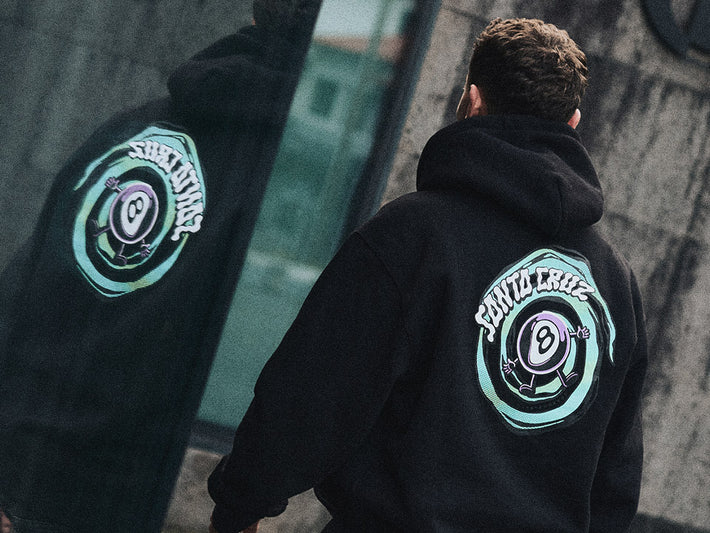 Santa Cruz Men's Clothing - a person wearing a black hoodie with a neon green logo on the back, standing against a blurred urban background.