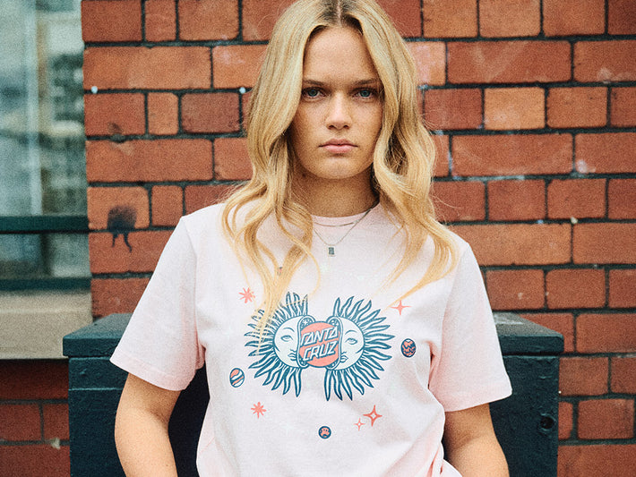 Santa Cruz Women's Clothing - a woman wearing a white t-shirt with a colorful graphic design against a brick wall.