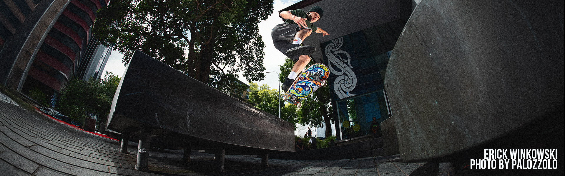 Santa Cruz Skateboards - a skateboarder performs a trick off a ledge in an urban setting with trees and buildings in the background. The image conveys action and excitement.
