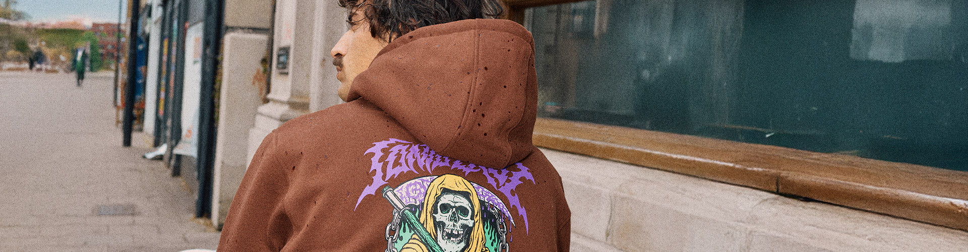 Santa Cruz Out of Time Graphic Collection. A person in a brown Santa Cruz hoodie with a colorful grim reaper graphic sits, with a city street in the background.