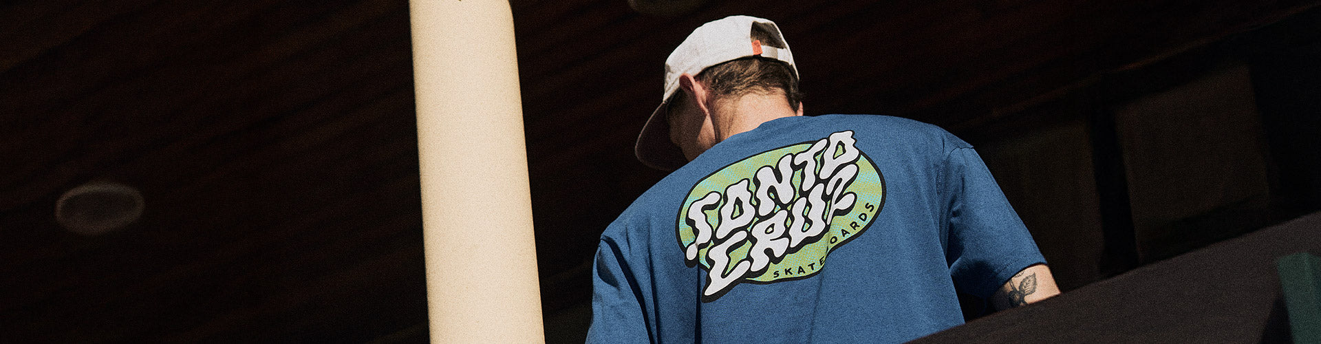 Santa Cruz Winkowski Vertigo Dot Collection. Man in a blue Santa Cruz t-shirt with text graphic and white cap stands outdoors, facing away. 