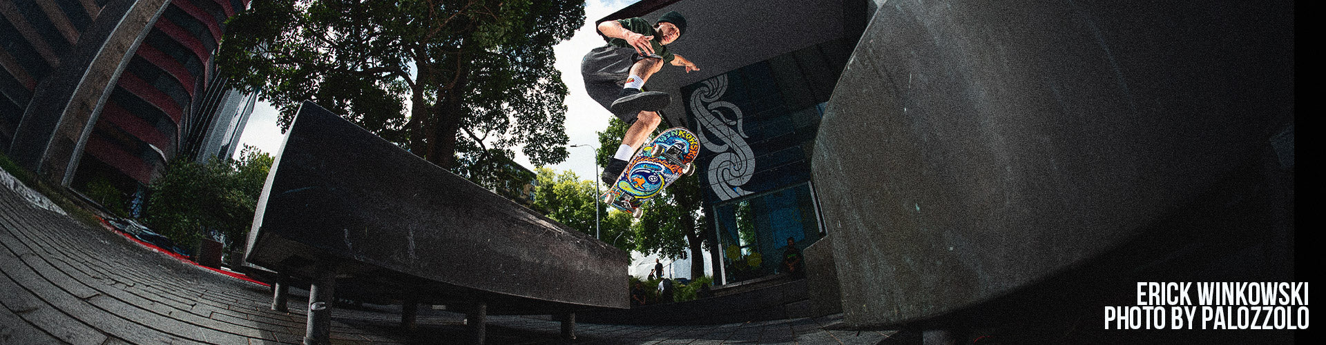 Santa Cruz Skateboards - a skateboarder performs a trick off a ledge in an urban setting with trees and buildings in the background. The image conveys action and excitement.