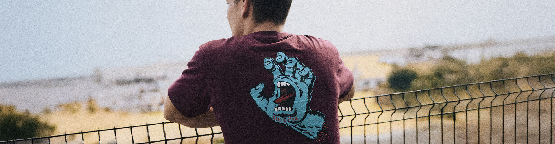 Santa Cruz Screaming Hand Collection. Man in a maroon Santa Cruz t-shirt featuring a screaming hand graphic on the back, leans on a fence.