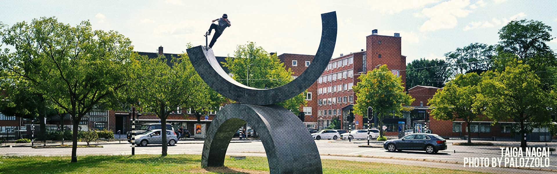 Santa Cruz skater performing a trick on a large abstract sculpture in an urban park with trees and buildings in the background