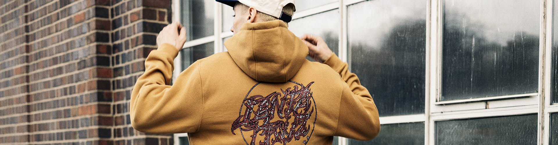 A person wearing a tan Santa Cruz Scourge hoodie with a graphic print on the back, standing in front of a brick wall and window.