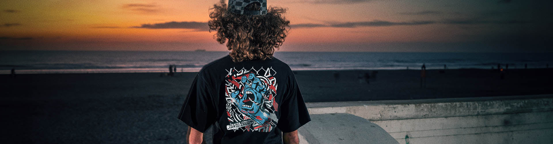 A person standing on a beach at sunset, viewed from behind, wearing a black Santa Cruz graphic T-shirt with a large damaged hand design on the back.