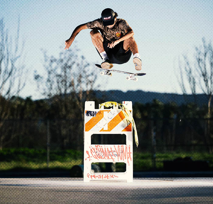 Santa Cruz skateboarder performing a trick above a traffic barrier with trees in the background