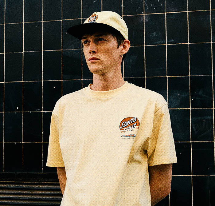 Man wearing a custard Santa Cruz t-shirt with a logo and a cap, standing against a black tiled wall.