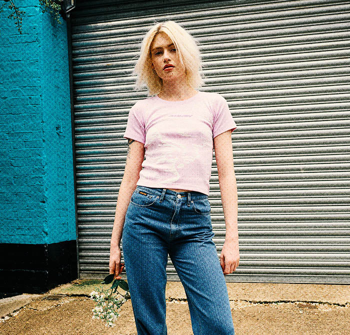Woman wearing a pink Santa Cruz t-shirt and blue jeans standing in front of a teal wall and metal shutter.