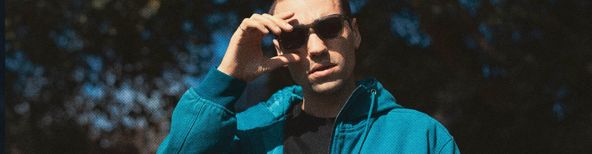 Man wearing a blue Santa Cruz jacket and Santa Cruz sunglasses with a blurred natural background.