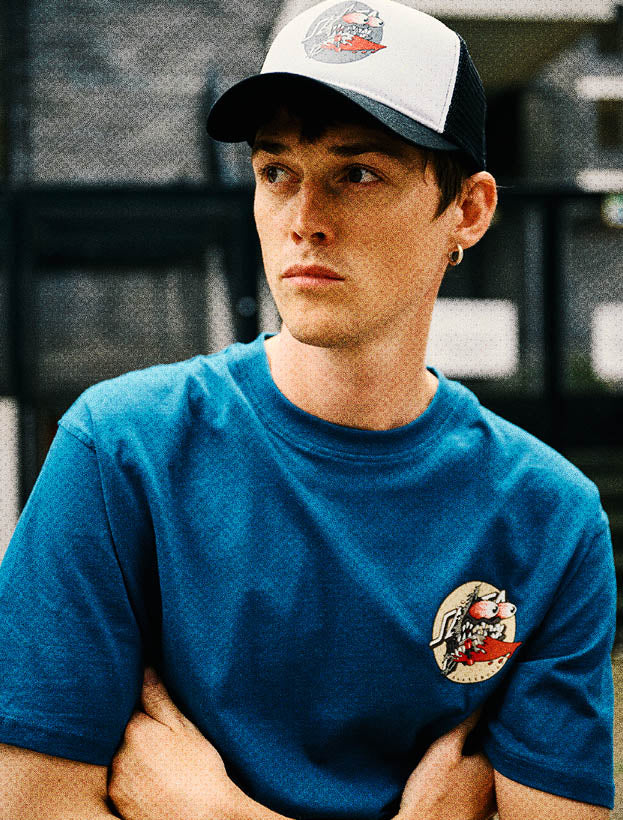 Man wearing a blue Santa Cruz t-shirt with a logo and a cap, standing against a blurred background.