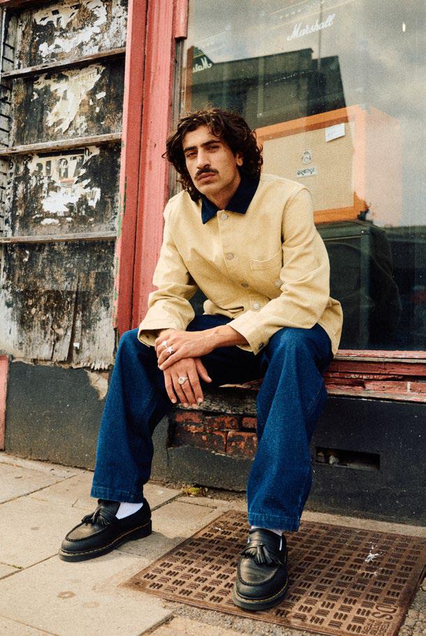 Man sitting on a bench wearing a yellow Santa Cruz Strike jacket, Kayce dark jeans and loafers with a store front in the background.