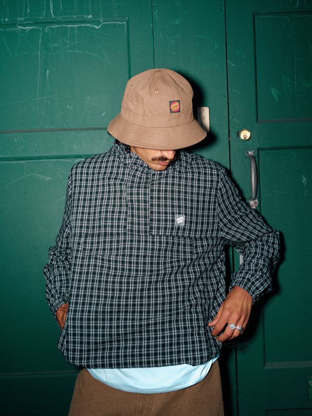 Person wearing a black check Santa Cruz Henri Jacket and beige bucket hat against a green door.