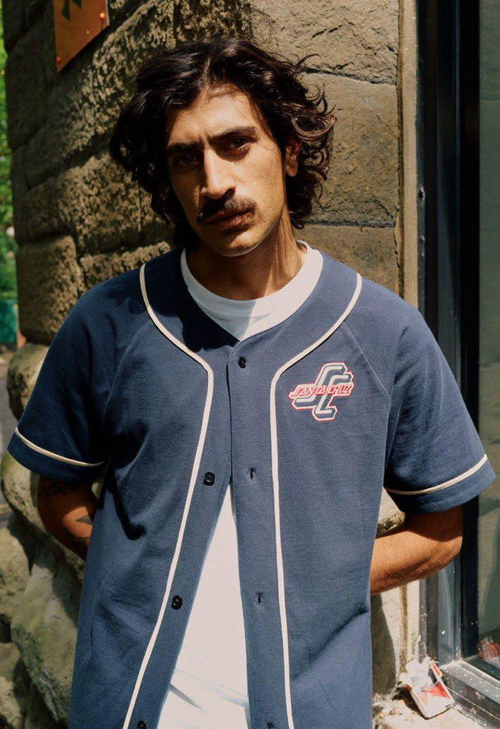 Man wearing a navy Santa Cruz Outer OGSC baseball shirt, standing against a stone wall.