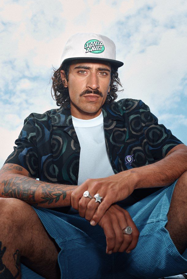 Man wearing a white Vertigo cap with a Santa Cruz logo, sitting against a cloudy sky.