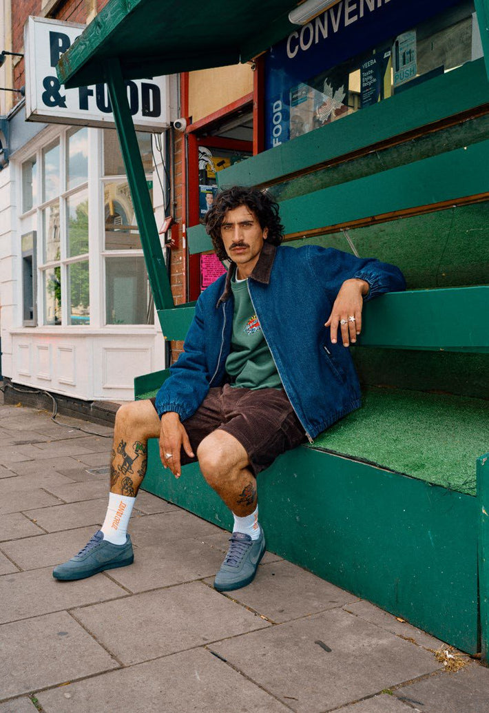 Man sitting on a green fruit stand wearing a Santa Cruz Kayce denim jacket and shorts.