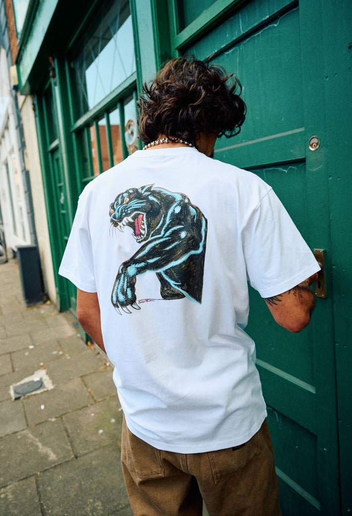 Person wearing a white Santa Cruz Natas Sketch t-shirt with a graphic of a panther on the back, standing in front of a green door.