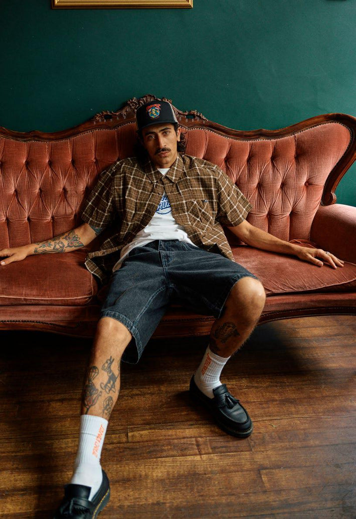 Man sitting on a vintage pink sofa wearing a Santa Cruz Eli brown check shirt, denim shorts, and black shoes.