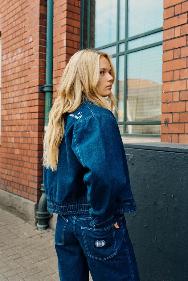 Woman wearing a blue denim Santa Cruz Split Sun jacket and shorts standing against a brick wall.