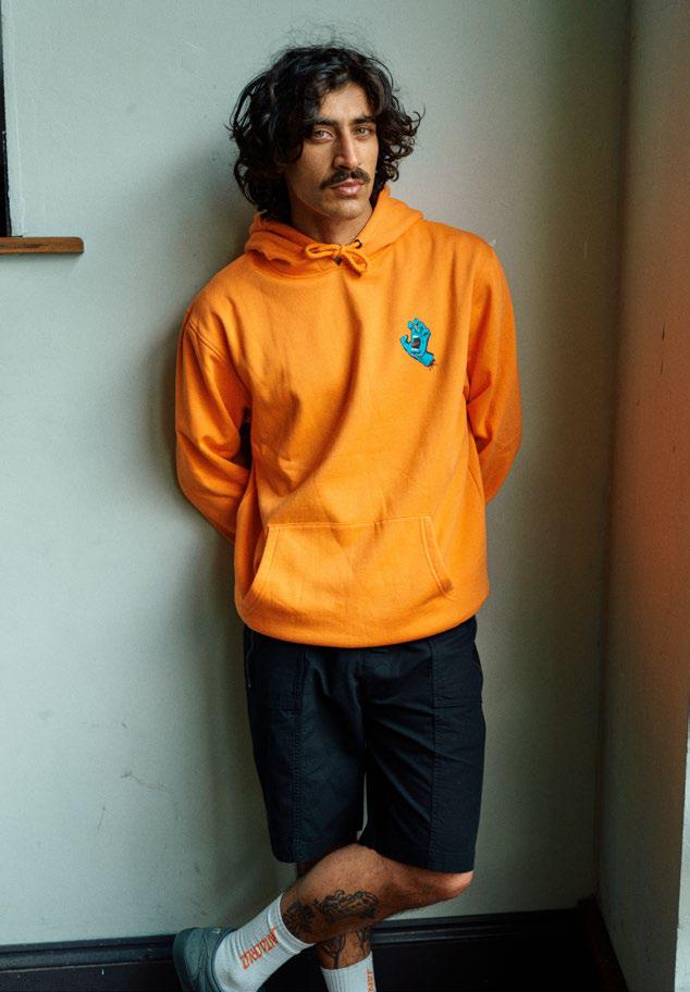Man wearing an orange Santa Cruz Screaming Hand hoodie and Clyde shorts, standing against a plain wall.